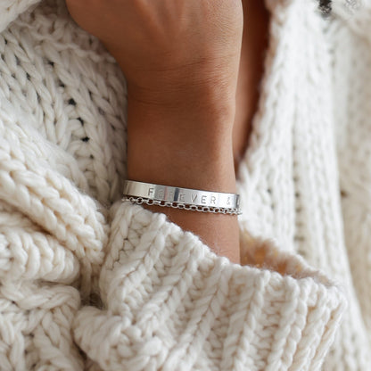 Close-up of a wrist wearing a silver bracelet with 'FEARLESS' engraved, against a textured white fabric background.