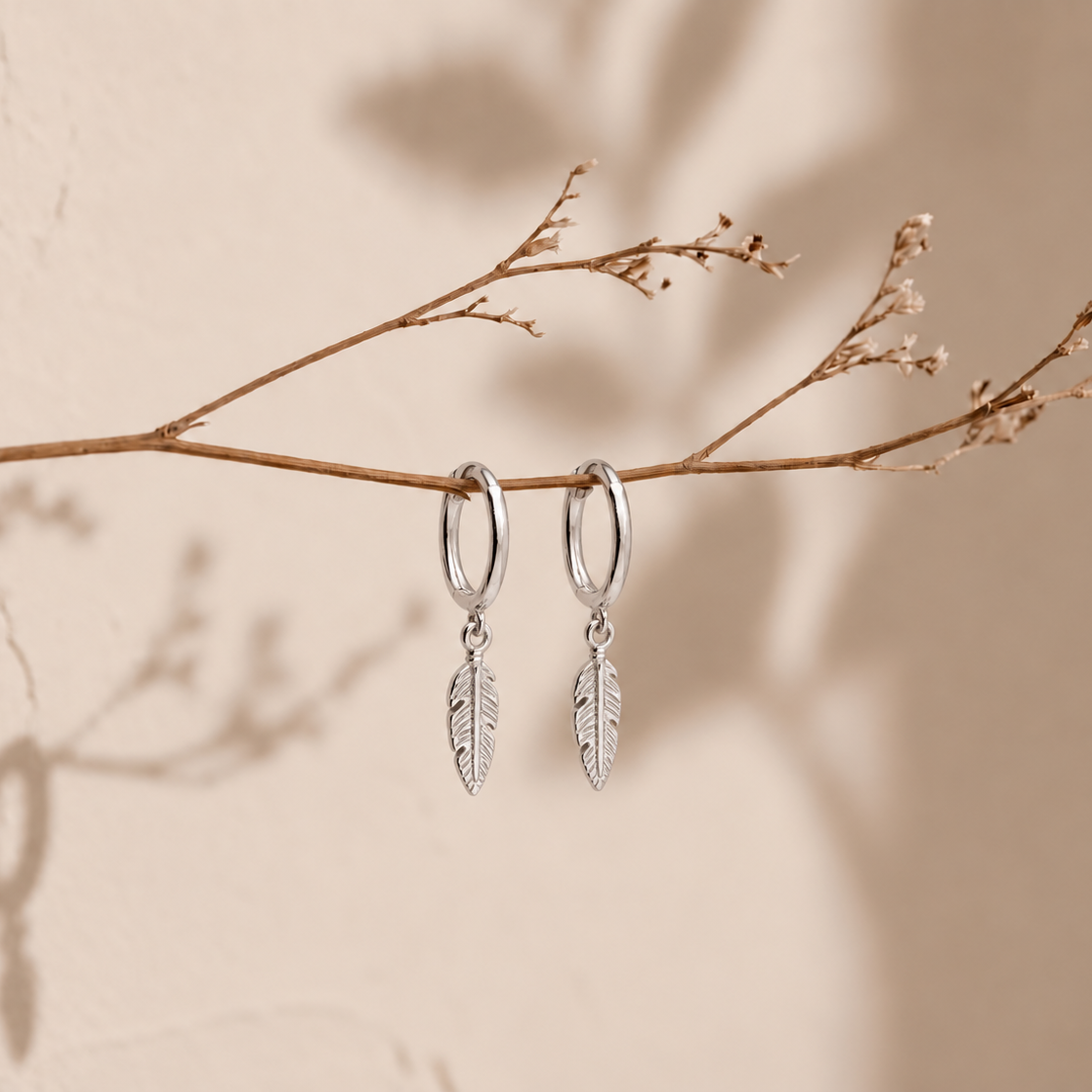 A pair of silver hoop earrings with dangling feather charms, hanging from a dried twig against a warm beige wall with soft shadows.