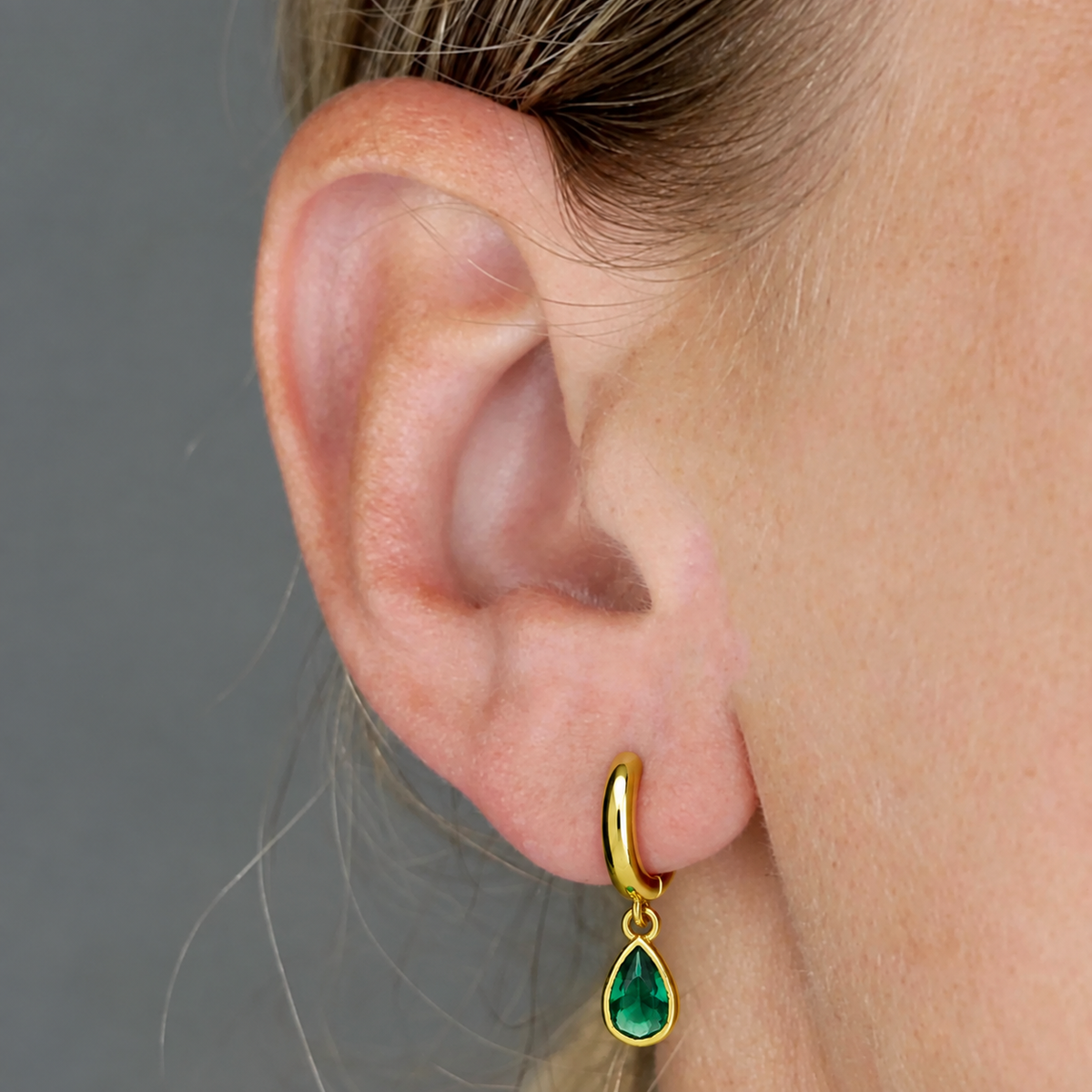 A close-up of a person's ear wearing a small gold huggie hoop earring with a emerald teardrop gemstone, against a plain gray background.