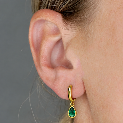 A close-up of a person's ear wearing a small gold huggie hoop earring with a emerald teardrop gemstone, against a plain gray background.
