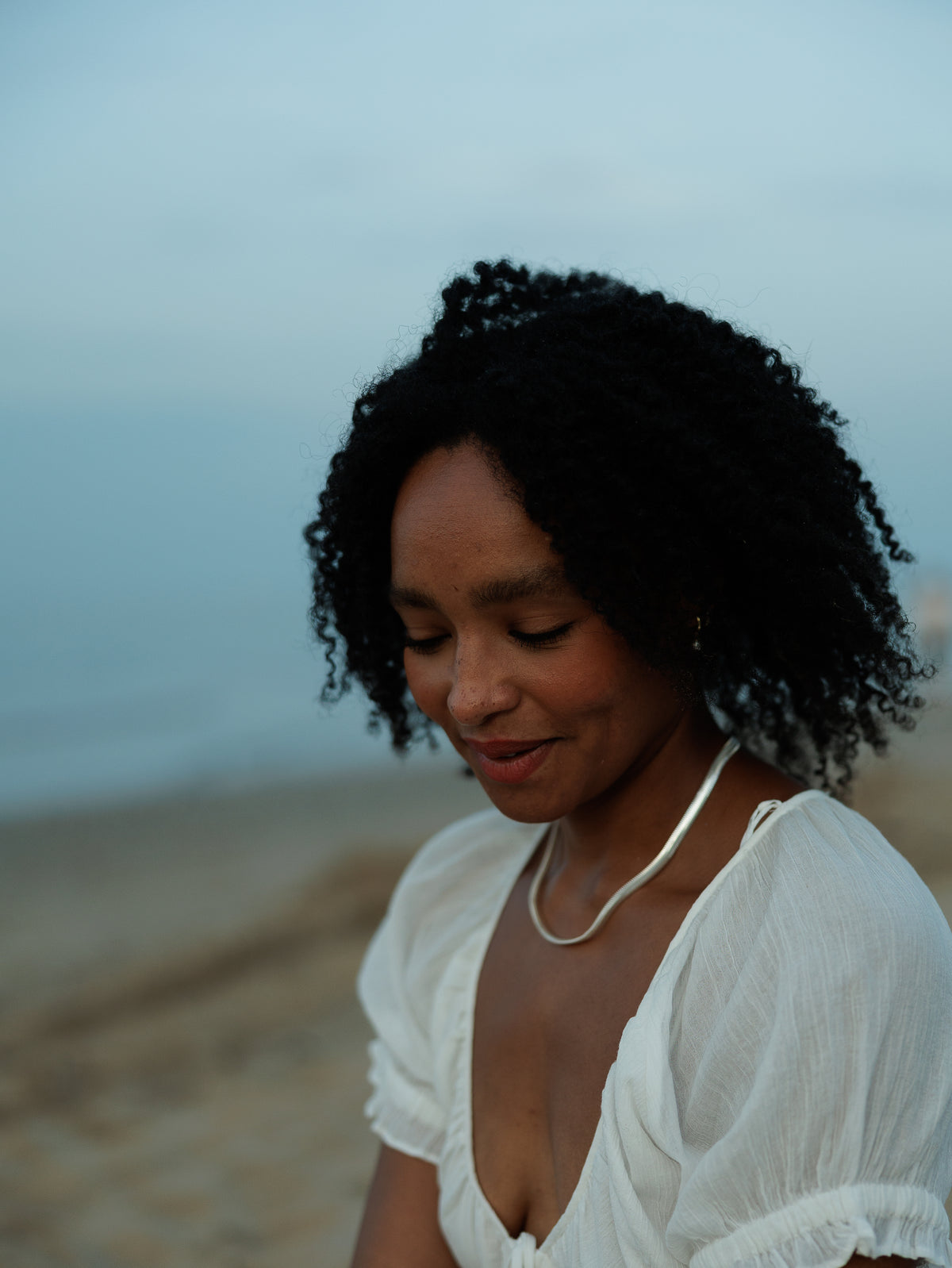 A woman with curly black hair wearing a white ruffled top and a silver necklace.