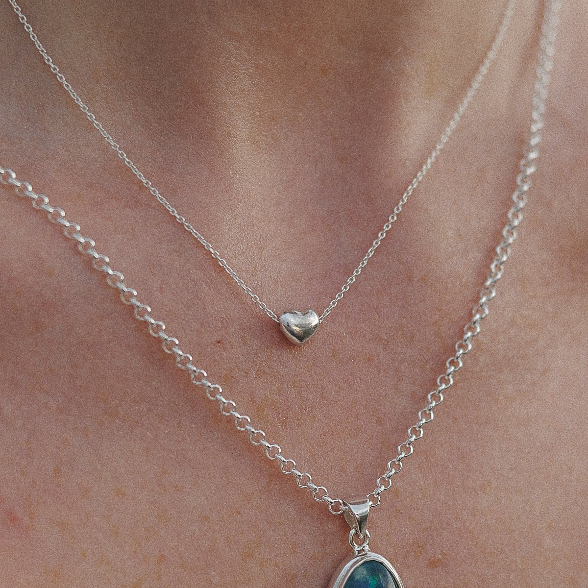 A close-up of a person's neck wearing two layered silver necklaces. The shorter necklace features a small polished heart pendant.
