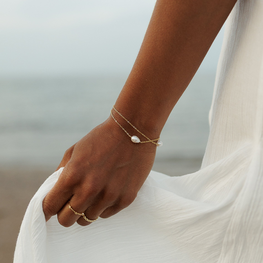 A close-up lifestyle shot of a person on a beach wearing delicate gold chain bracelets and rings, holding the hem of a white dress.