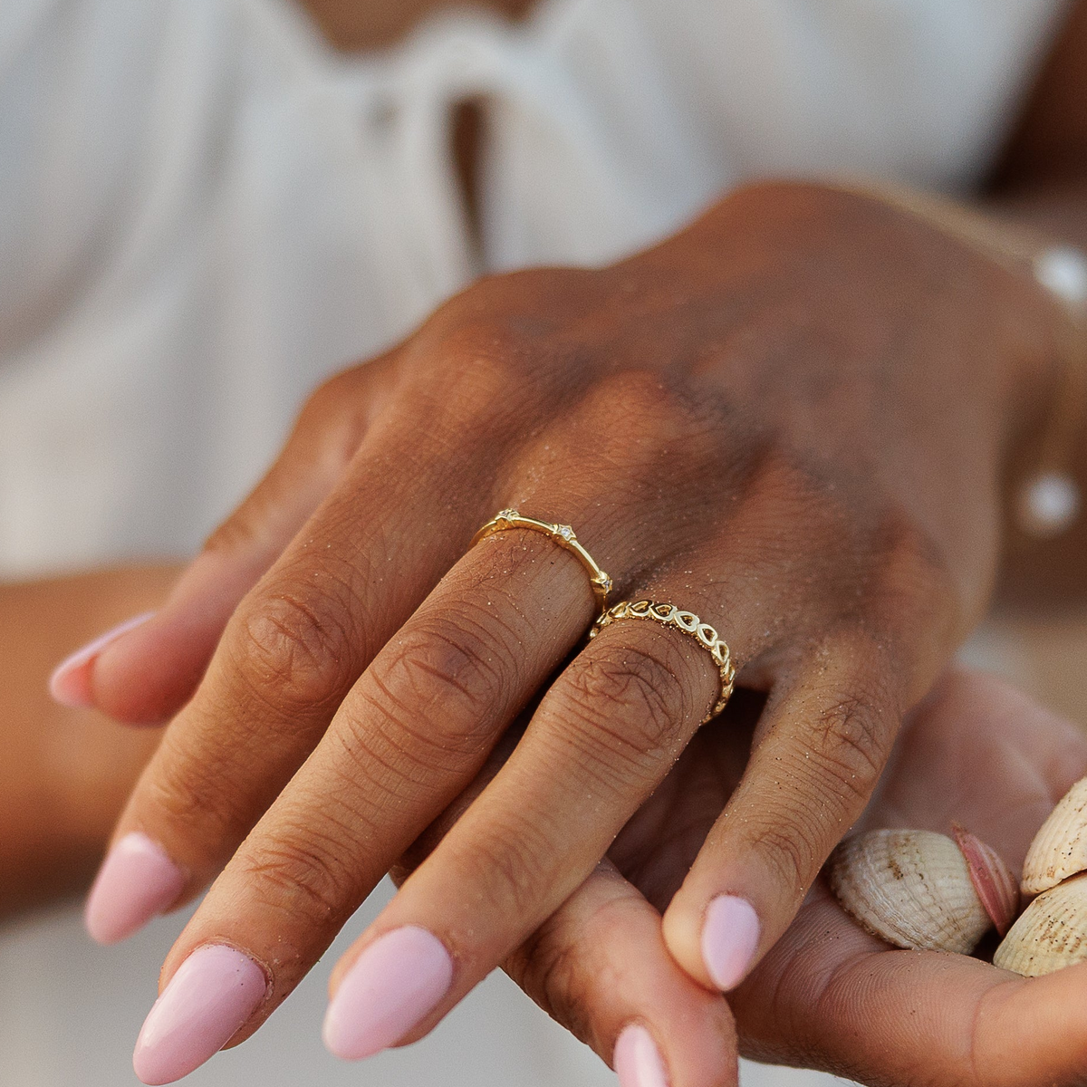 A close-up of a person's hands holding small seashells, wearing a white dress and gold rings, with a soft beige background.