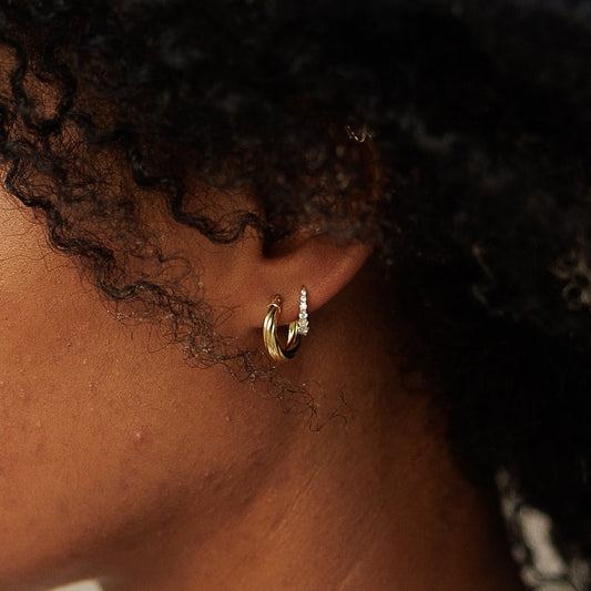A woman with voluminous, curly black hair is shown in profile against a soft-focus beach background. She is wearing a white top and a delicate gold necklace.