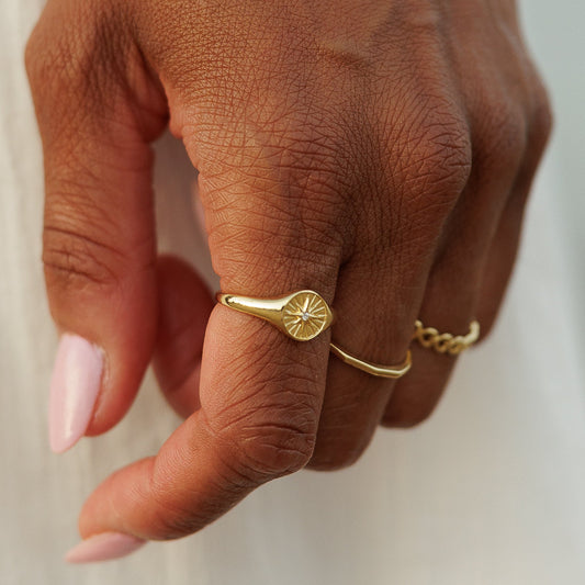 A close-up of a person's hand wearing delicate gold jewelry, including a layered chain bracelet and small rings, against a soft white dress and blurred outdoor background.
