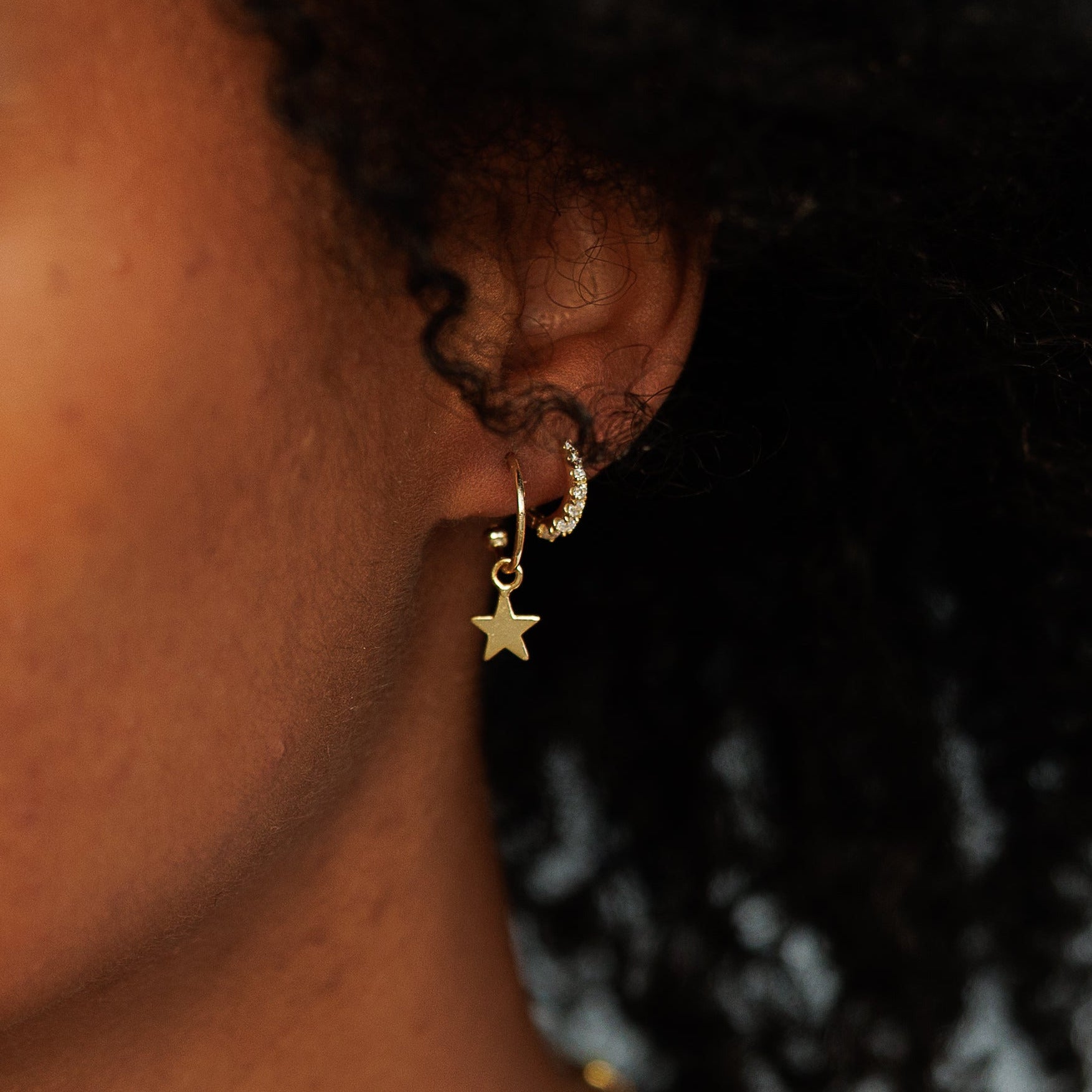 A close-up portrait of a woman with curly black hair wearing a delicate gold necklace and a small gold star earring, set against a soft, blurred outdoor background.