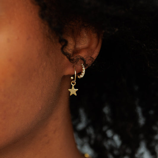 A close-up portrait of a woman with curly black hair wearing a delicate gold necklace and a small gold star earring, set against a soft, blurred outdoor background.