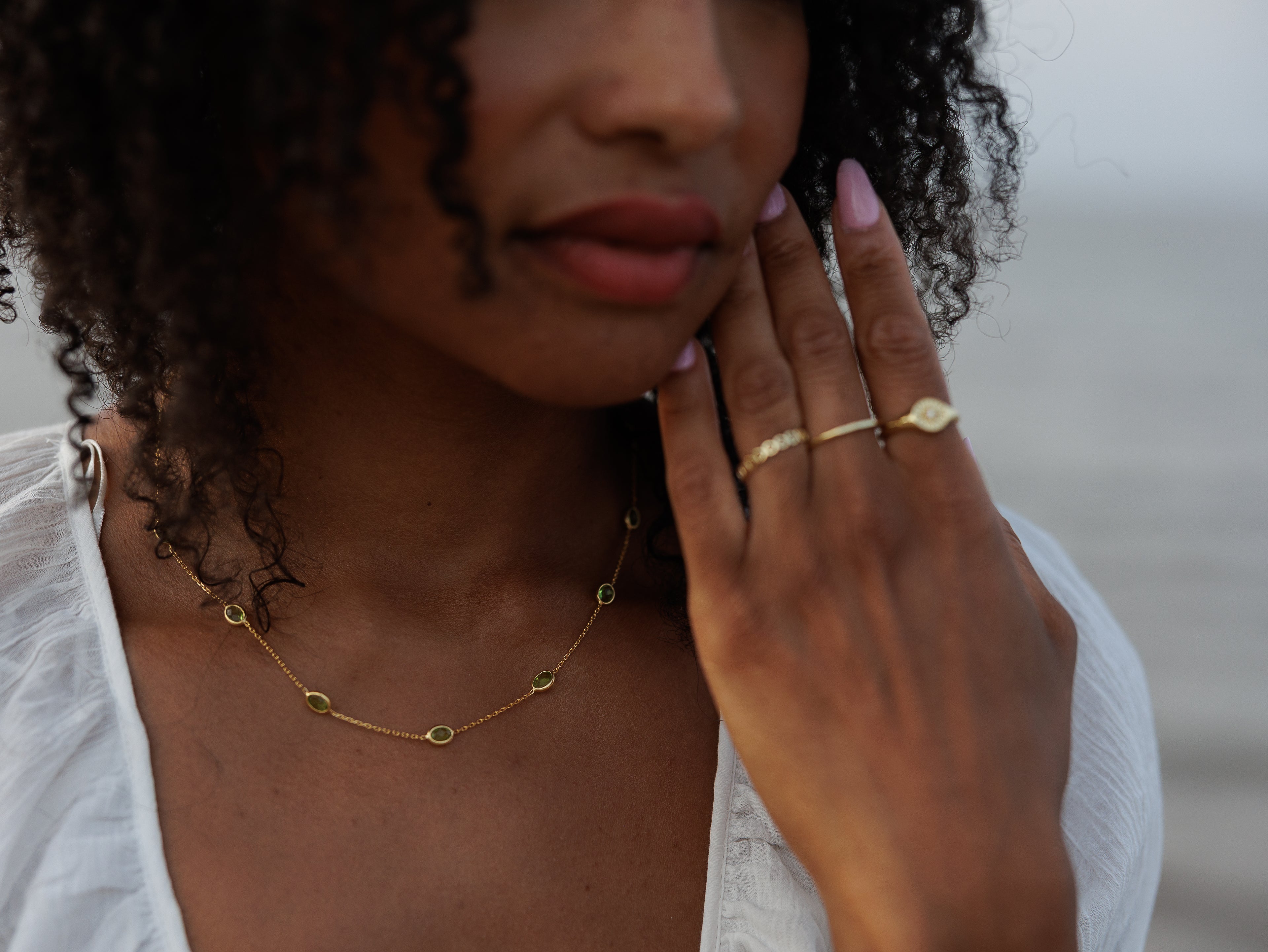 A close-up lifestyle shot of a woman wearing a delicate gold necklace with small circular charms and matching gold rings on her fingers, set against a soft, blurred background.