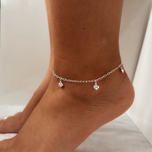 Silver anklet with heart-shaped charms on a person's ankle against a neutral background