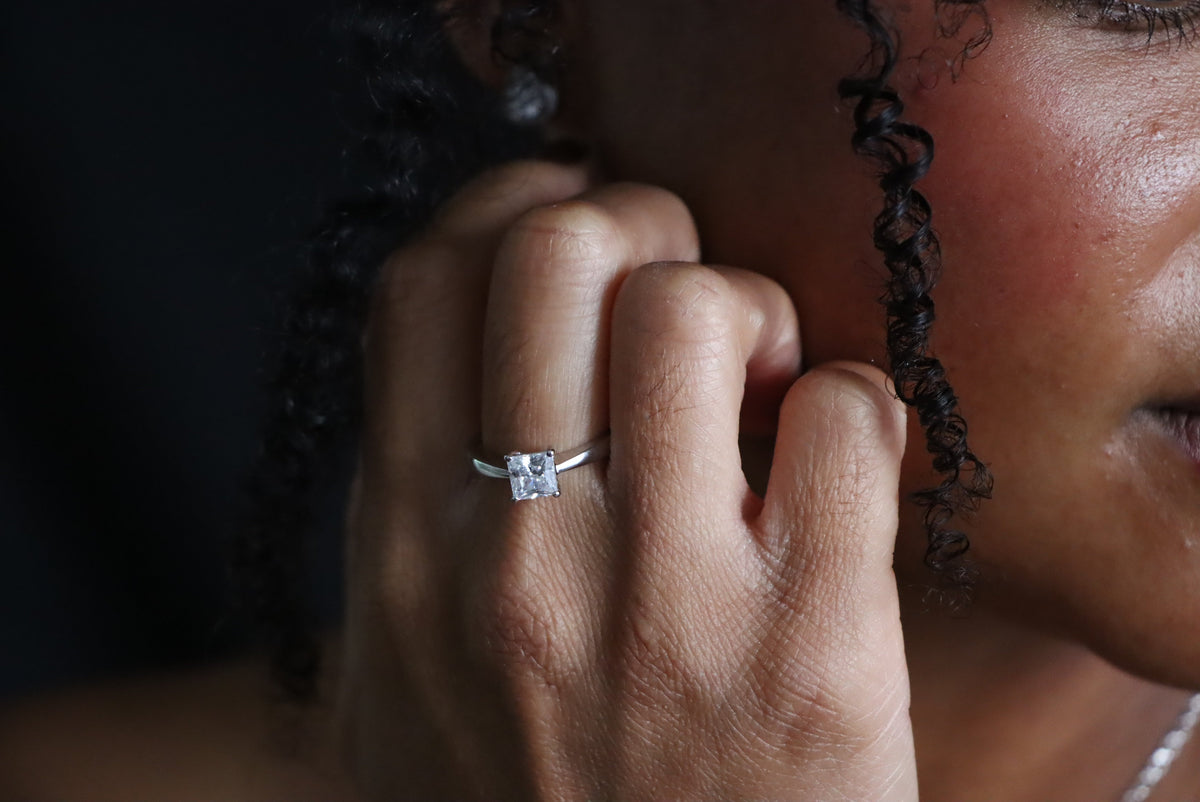 Sterling silver ring with a square princess-cut clear stone, worn on a hand beside the model’s face