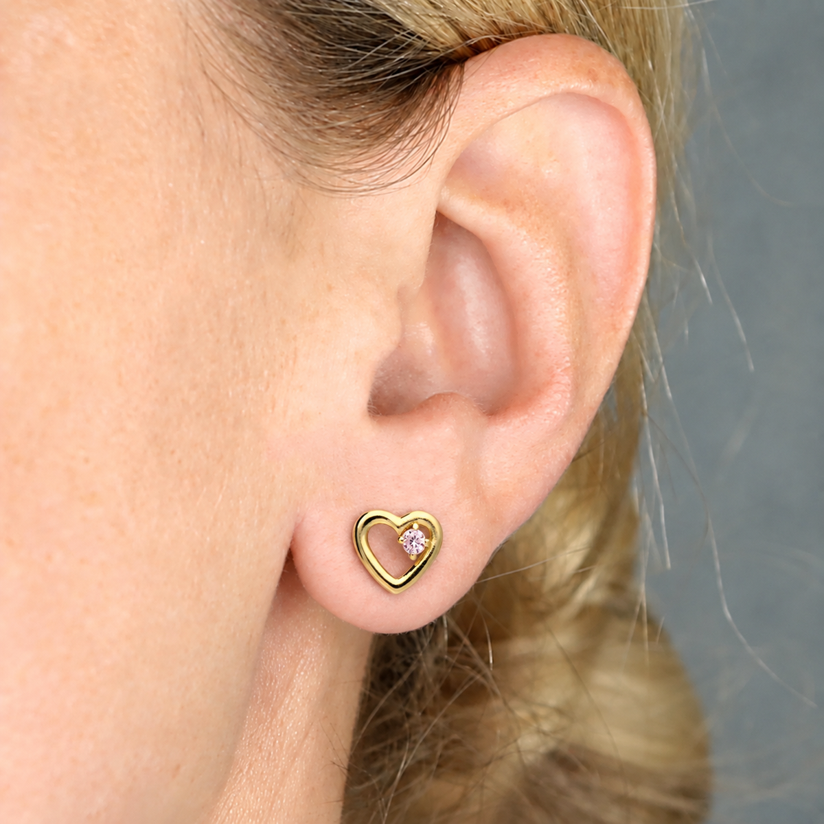A close-up of a person's ear wearing a small gold heart-shaped stud earring with a pink crystal stone