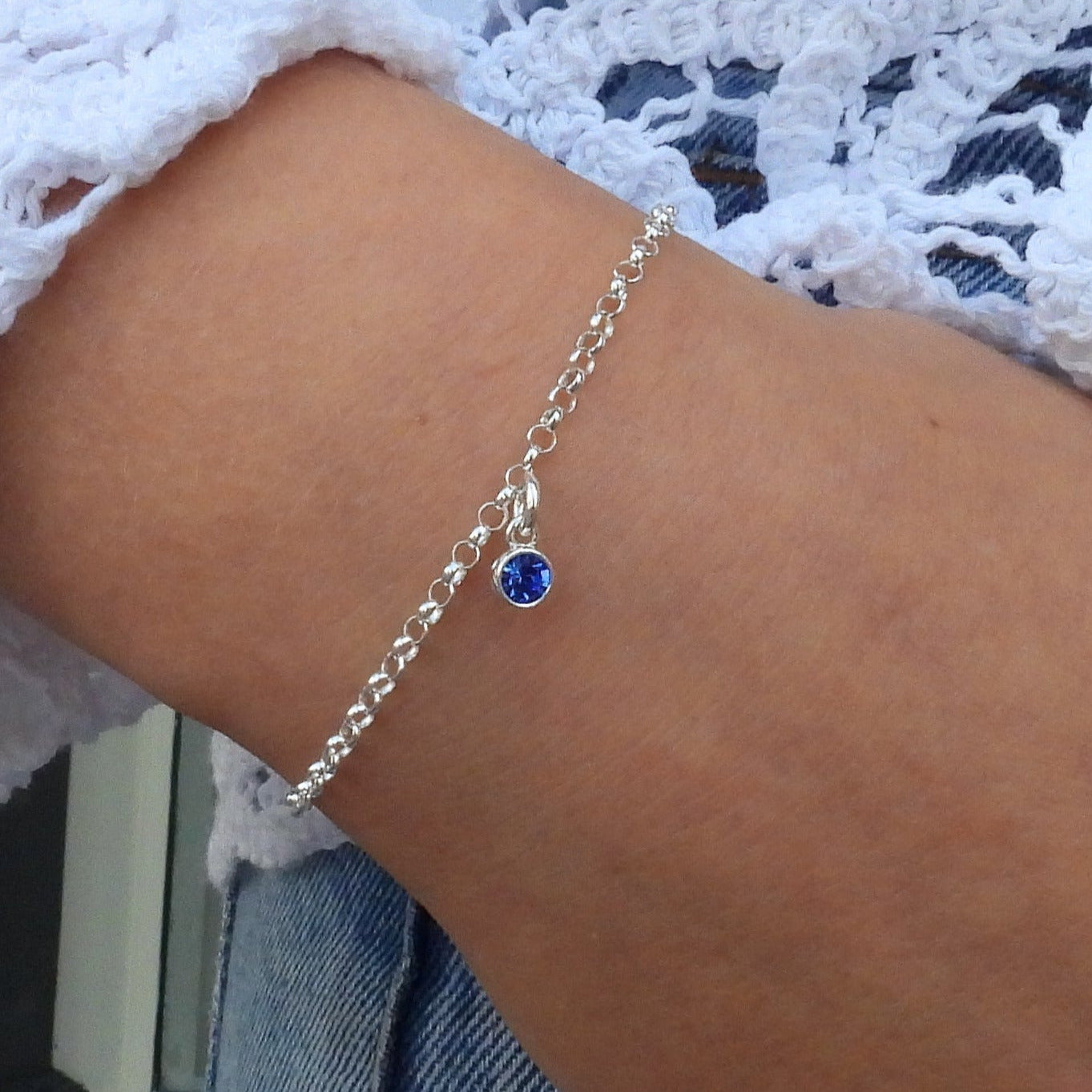 Close-up of a hand wearing a silver bracelet and ring with blue stones, against a white lace garment and denim background.