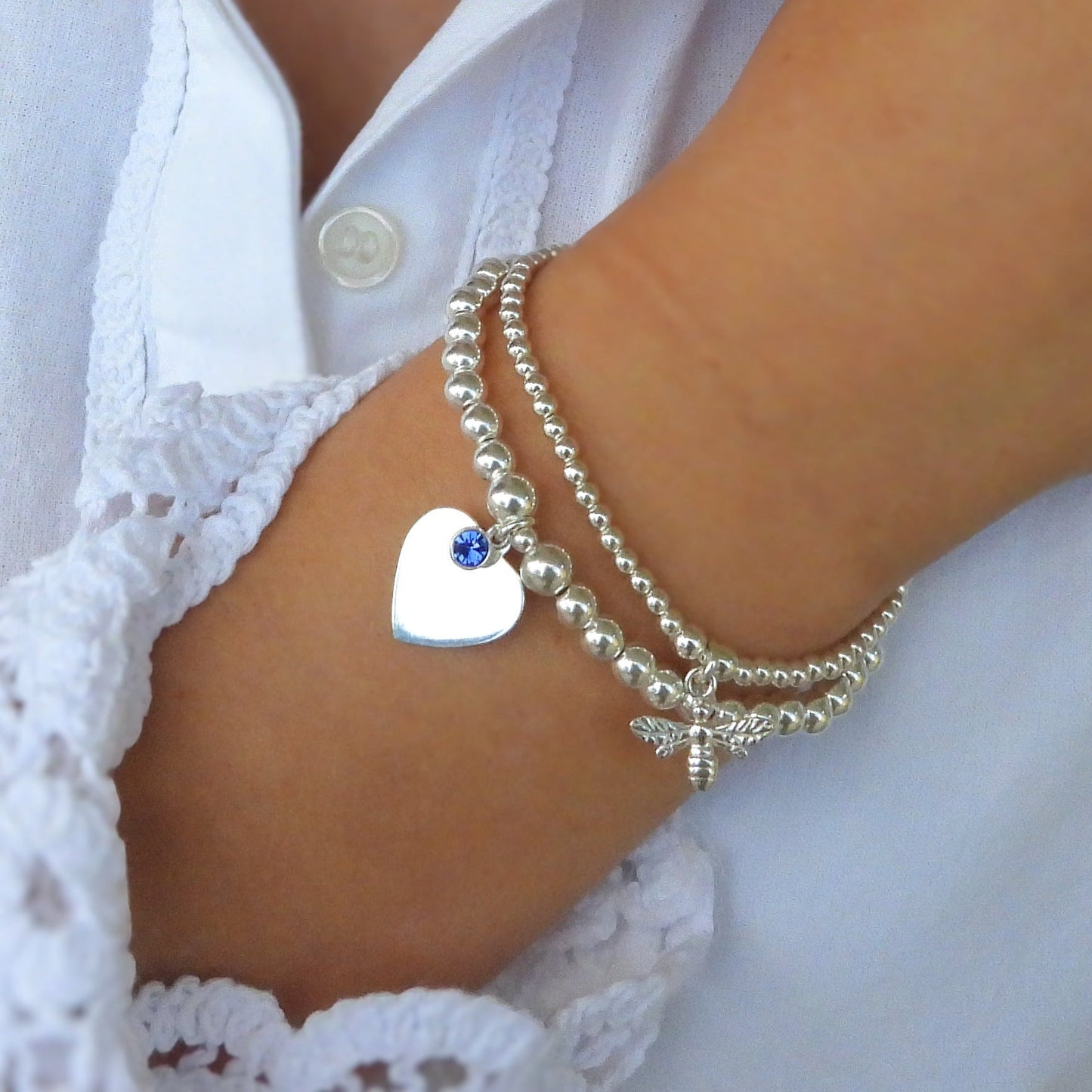 Close-up of a person wearing a silver bracelet with a heart charm and a necklace with a blue pendant on a white lace shirt.