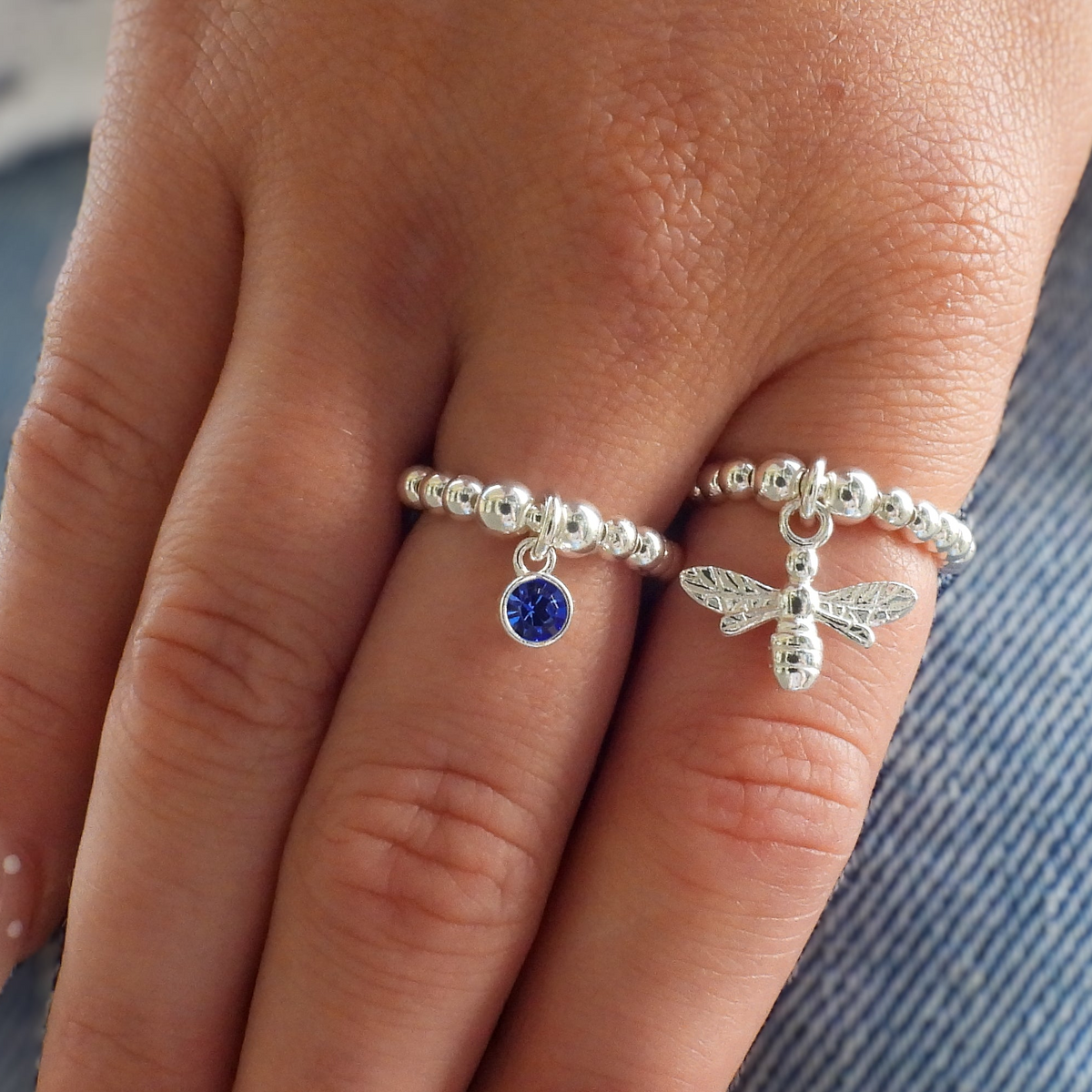 Two silver rings worn on a hand, one featuring a small round blue gemstone and the other featuring a detailed bee charm, set against a background of blue denim.