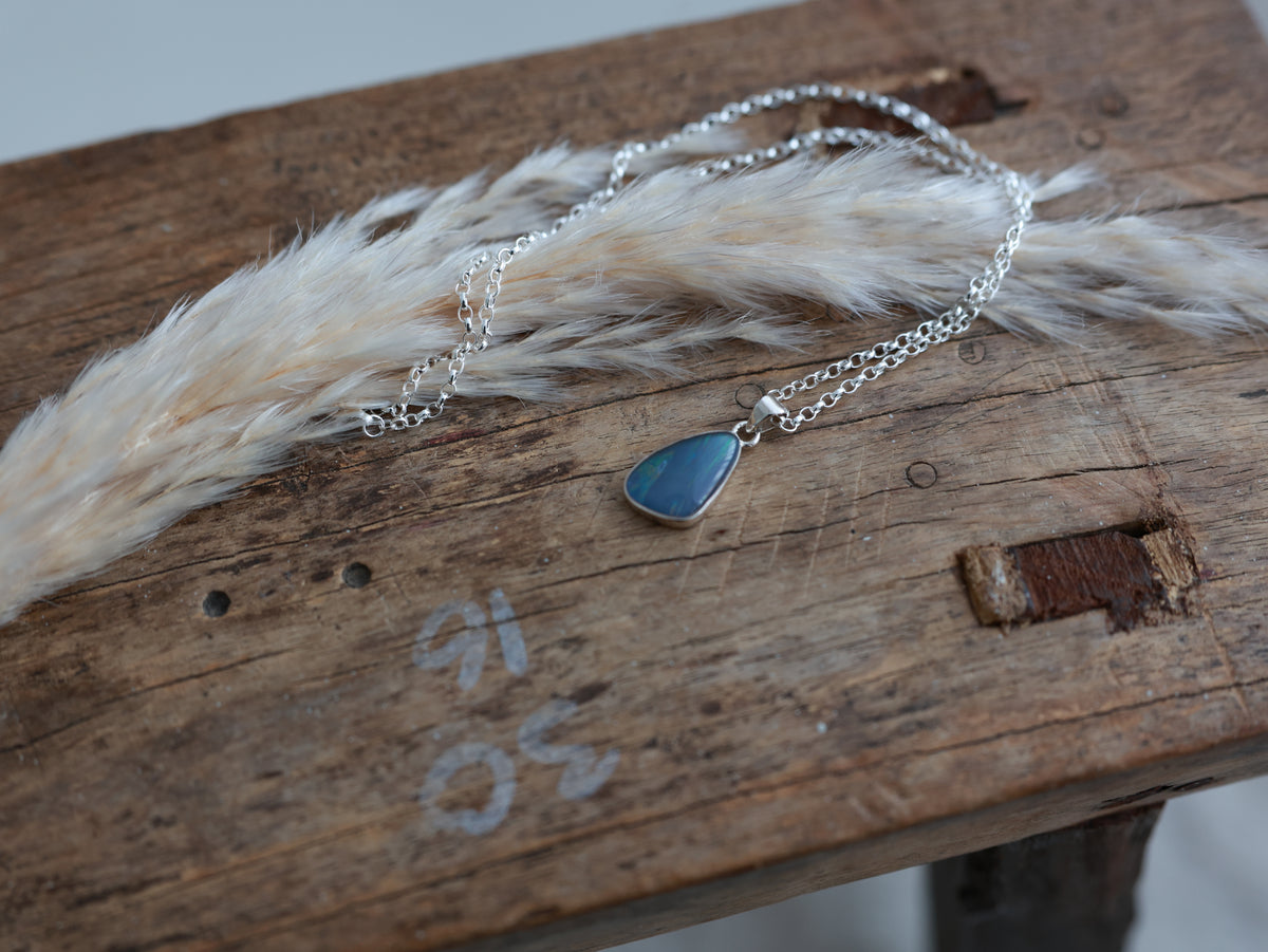 Sterling silver Australian Opal Treasure Necklace with a blue freeform pendant, displayed on rustic wood beside white dried foliage
