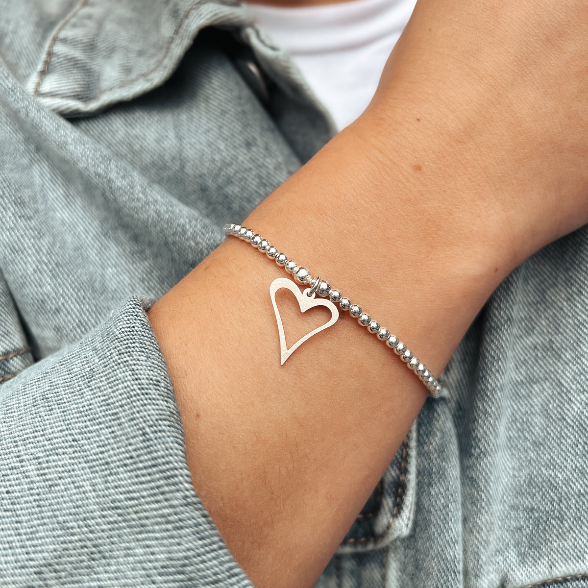 Sterling silver beaded bracelet with an open heart charm, worn on a wrist beside a denim sleeve