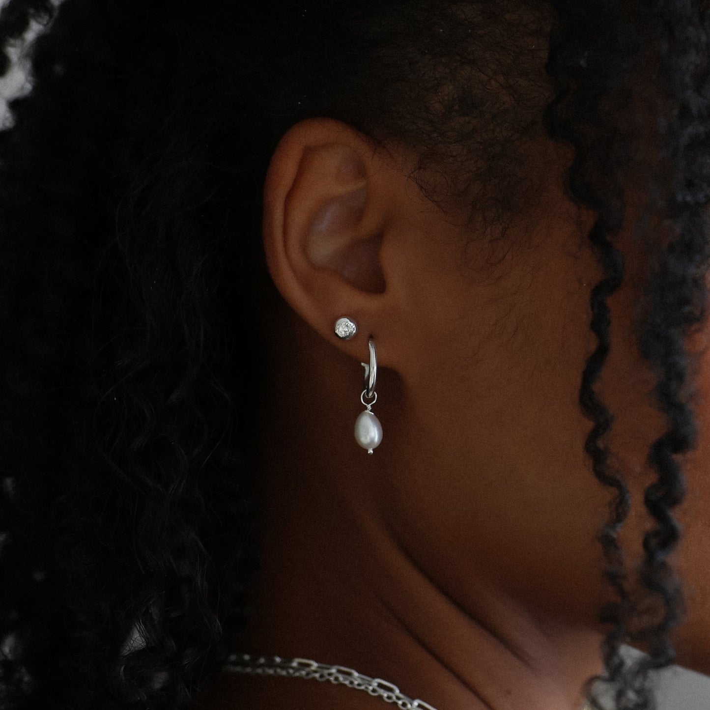 Profile of a young girl with braided hair wearing pearl earrings and a necklace, with a blurred background.