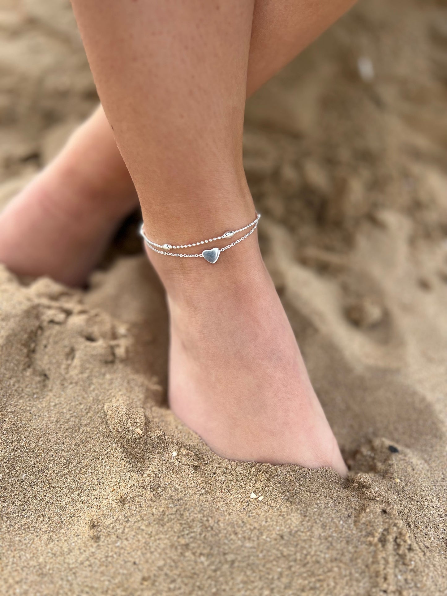 Close-up of a person's ankle with double chain heart anklet on, on a sandy background