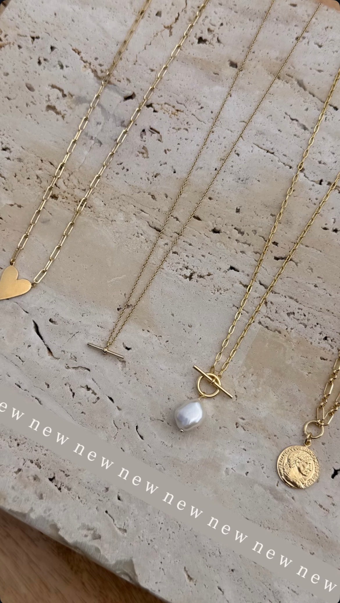 Gold necklaces on a pale stone surface, including a heart pendant, dainty T-bar, freshwater pearl T-bar and coin pendant