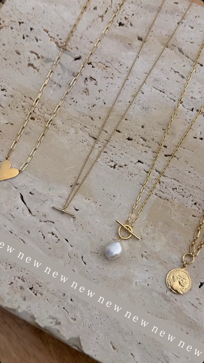 Gold necklaces on a pale stone surface, including a heart pendant, dainty T-bar, freshwater pearl T-bar and coin pendant
