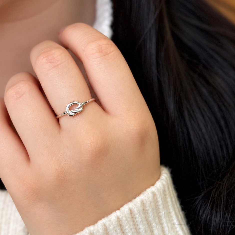A close-up of a person's hand wearing a delicate silver ring with a small open circular design, paired with a cream-colored ribbed knit sweater.