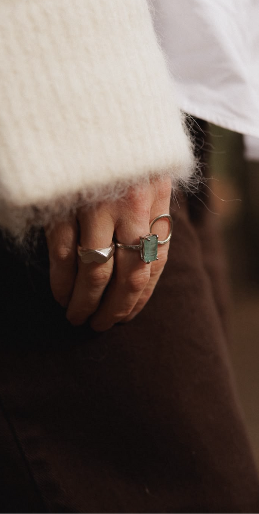 Close-up of a person's hand wearing multiple rings, including a green kyanite rectangular ring styled with a white fuzzy sleeve and dark brown clothing.