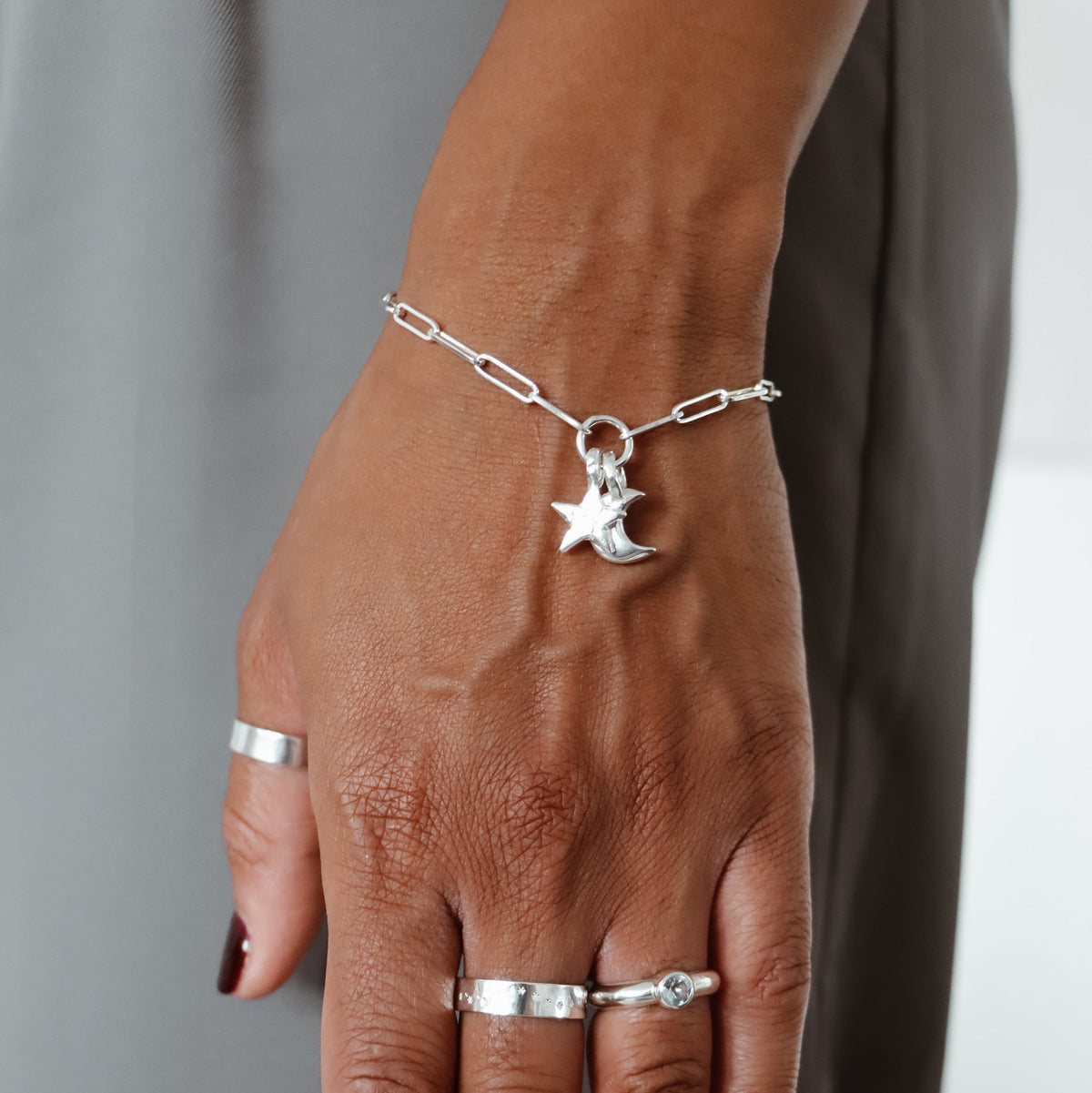 A close-up of a person's hand wearing a delicate silver chain bracelet with a crescent moon and star charm, along with several silver rings, against a neutral gray background.