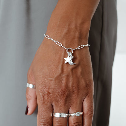 A close-up of a person's hand wearing a delicate silver chain bracelet with a crescent moon and star charm, along with several silver rings, against a neutral gray background.