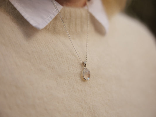 A close-up lifestyle shot of a delicate silver chain necklace with a teardrop-shaped moonstone pendant, worn by a person in a cream-colored knit sweater over a white collared shirt.