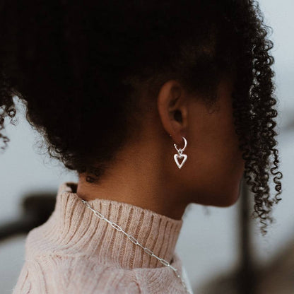 Sterling silver open heart charm hanging from a small hoop earring, worn by a model in a beige knitted jumper.