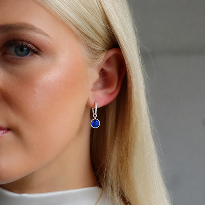 A close-up of a blonde woman wearing a silver drop earring with a round blue sapphire gemstone.