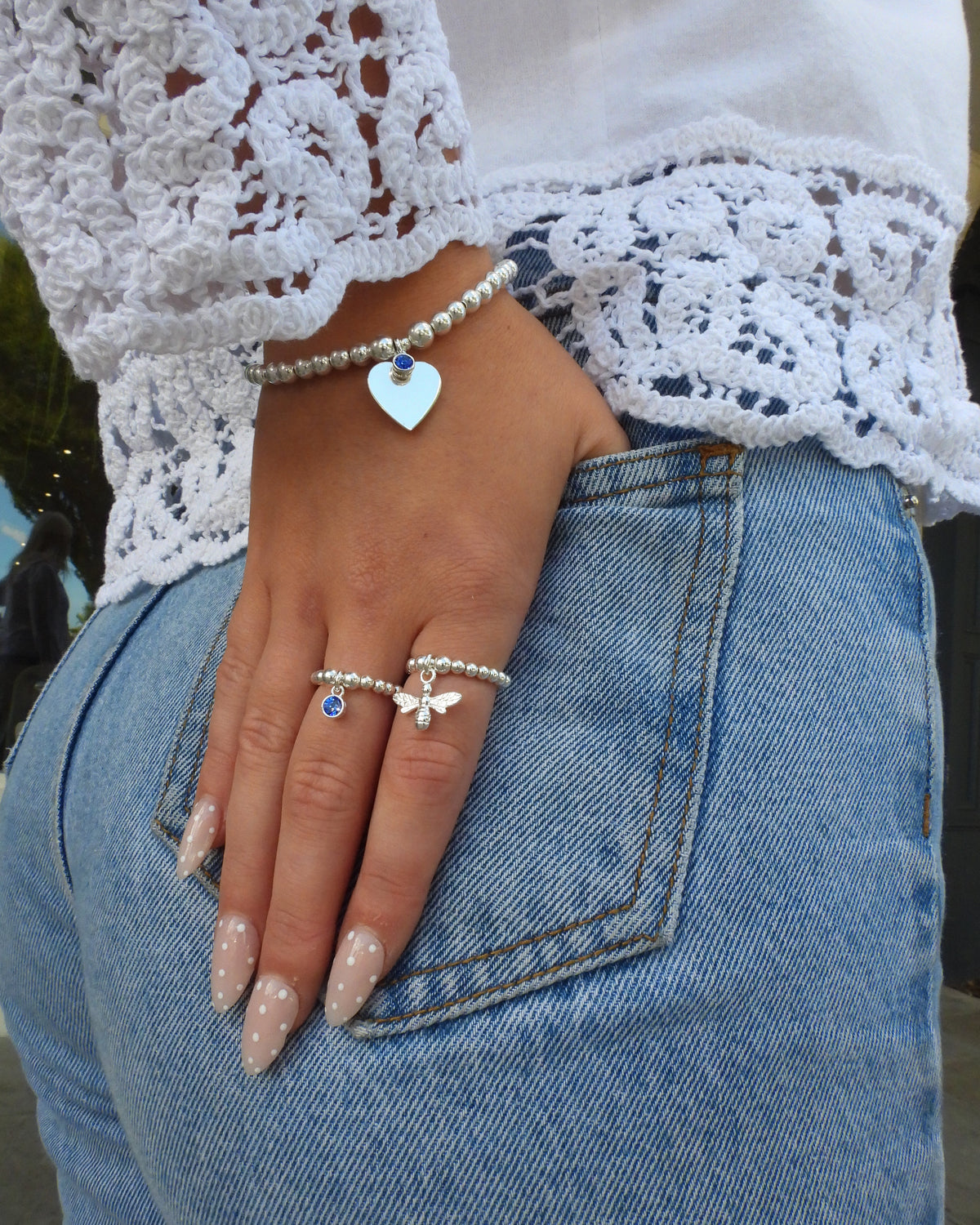 Sterling silver heart birthstone bracelet with a blue charm, worn with beaded rings against denim and white lace
