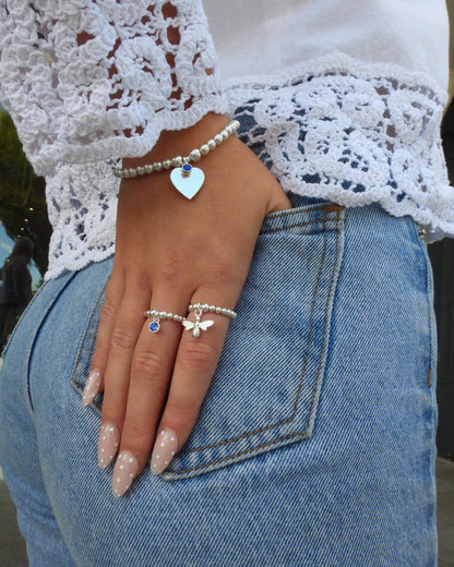 Sterling silver heart birthstone bracelet with a blue charm, worn with beaded rings against denim and white lace