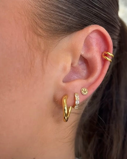 14ct gold-plated smiley face stud worn on an ear alongside hoop earrings and a gold ear cuff