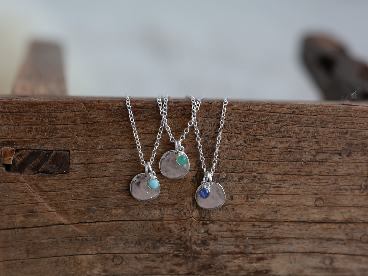 Three silver talisman necklaces with round disc pendants and pale blue, green and dark blue gemstone charms on rustic wood