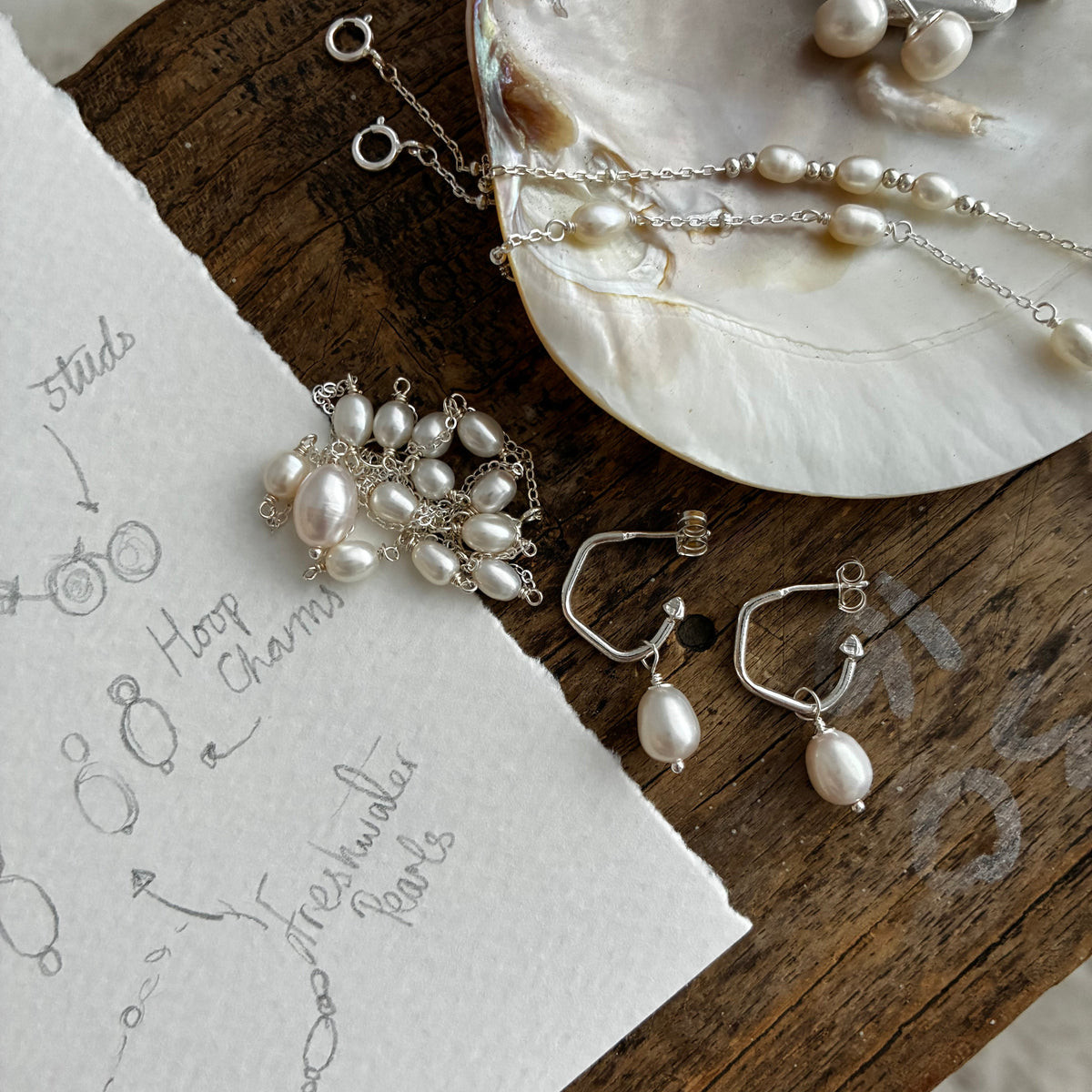 Silver jewelry with freshwater pearls arranged on a rustic wooden surface next to a piece of paper with hand-drawn sketches. The jewelry includes hoop earrings, a pendant, and chain pieces with silver hooks and dangling pearl drops.
