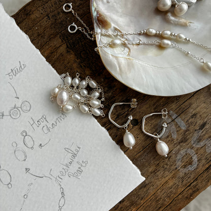 Silver jewelry with freshwater pearls arranged on a rustic wooden surface next to a piece of paper with hand-drawn sketches. The jewelry includes hoop earrings, a pendant, and chain pieces with silver hooks and dangling pearl drops.