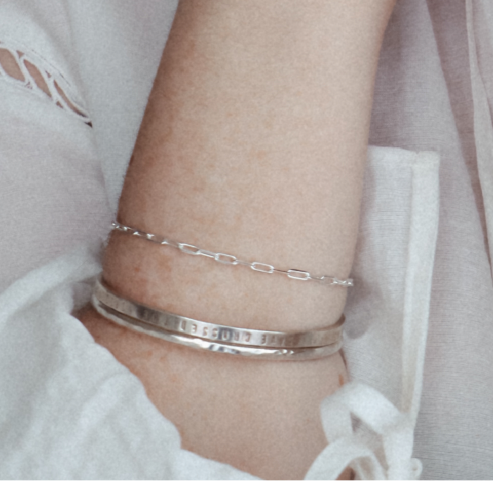 Sterling silver Teeny Trace Bracelet layered with two silver bangles on a wrist beside a white blouse.