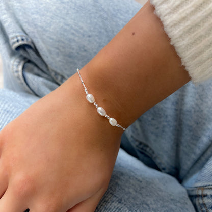 Silver-coloured fine chain bracelet with three white oval pearls, worn on a wrist against denim beside a cream sleeve